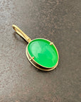 An oval pendant featuring a vibrant green chrysoprase gemstone in a gold bezel setting with a gold hook, displayed on a textured dark gray background.