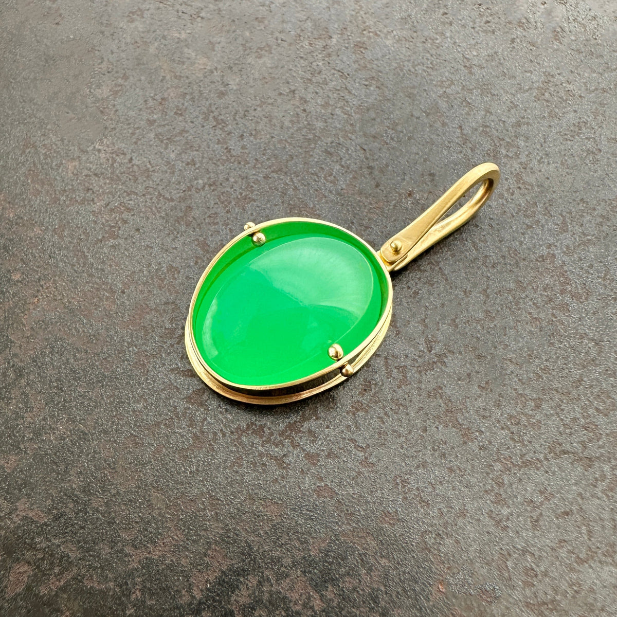 An oval pendant featuring a vibrant green chrysoprase gemstone in a gold bezel setting with a gold hook, displayed on a textured dark gray background.