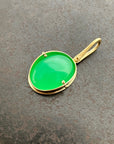 An oval pendant featuring a vibrant green chrysoprase gemstone in a gold bezel setting with a gold hook, displayed on a textured dark gray background.