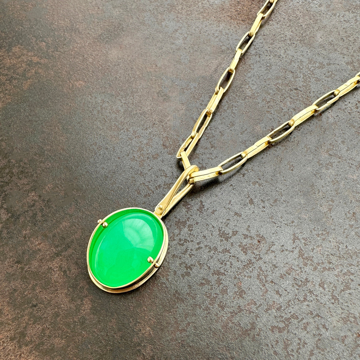 An oval pendant featuring a vibrant green chrysoprase gemstone in a gold bezel setting with a gold hook, displayed on a textured dark gray background.