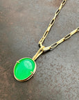 An oval pendant featuring a vibrant green chrysoprase gemstone in a gold bezel setting with a gold hook, displayed on a textured dark gray background.