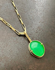 An oval pendant featuring a vibrant green chrysoprase gemstone in a gold bezel setting with a gold hook, displayed on a textured dark gray background.