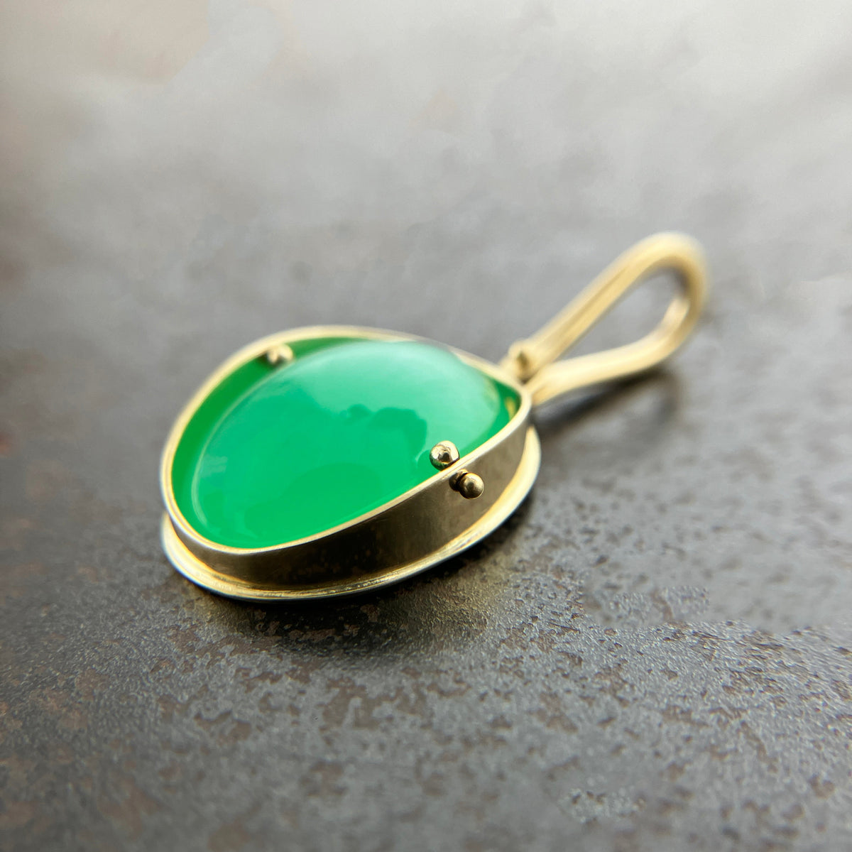 An oval pendant featuring a vibrant green chrysoprase gemstone in a gold bezel setting with a gold hook, displayed on a textured dark gray background.
