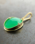 An oval pendant featuring a vibrant green chrysoprase gemstone in a gold bezel setting with a gold hook, displayed on a textured dark gray background.