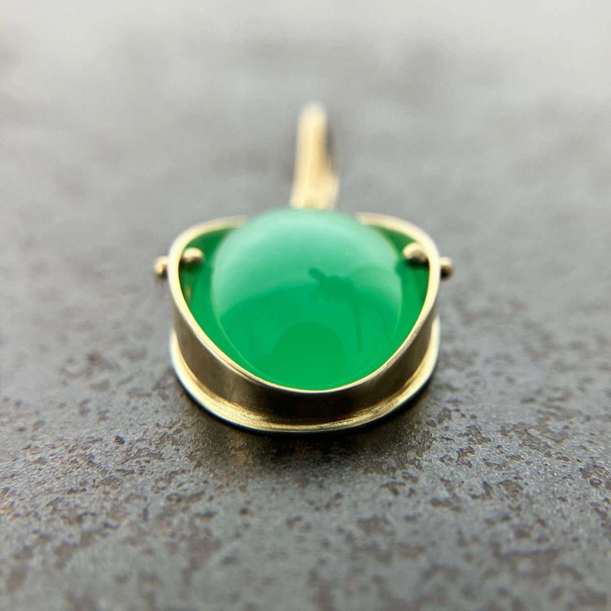 An oval pendant featuring a vibrant green chrysoprase gemstone in a gold bezel setting with a gold hook, displayed on a textured dark gray background.