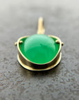 An oval pendant featuring a vibrant green chrysoprase gemstone in a gold bezel setting with a gold hook, displayed on a textured dark gray background.