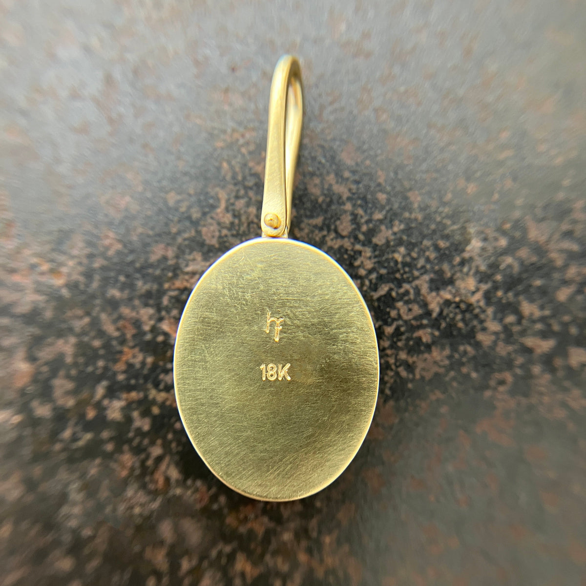 A close-up of a gold oval pendant with a loop at the top, showing a brushed finish and engraved '18K' and 'F' mark on the reverse side, set against a textured dark brown background.