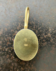 A close-up of a gold oval pendant with a loop at the top, showing a brushed finish and engraved '18K' and 'F' mark on the reverse side, set against a textured dark brown background.