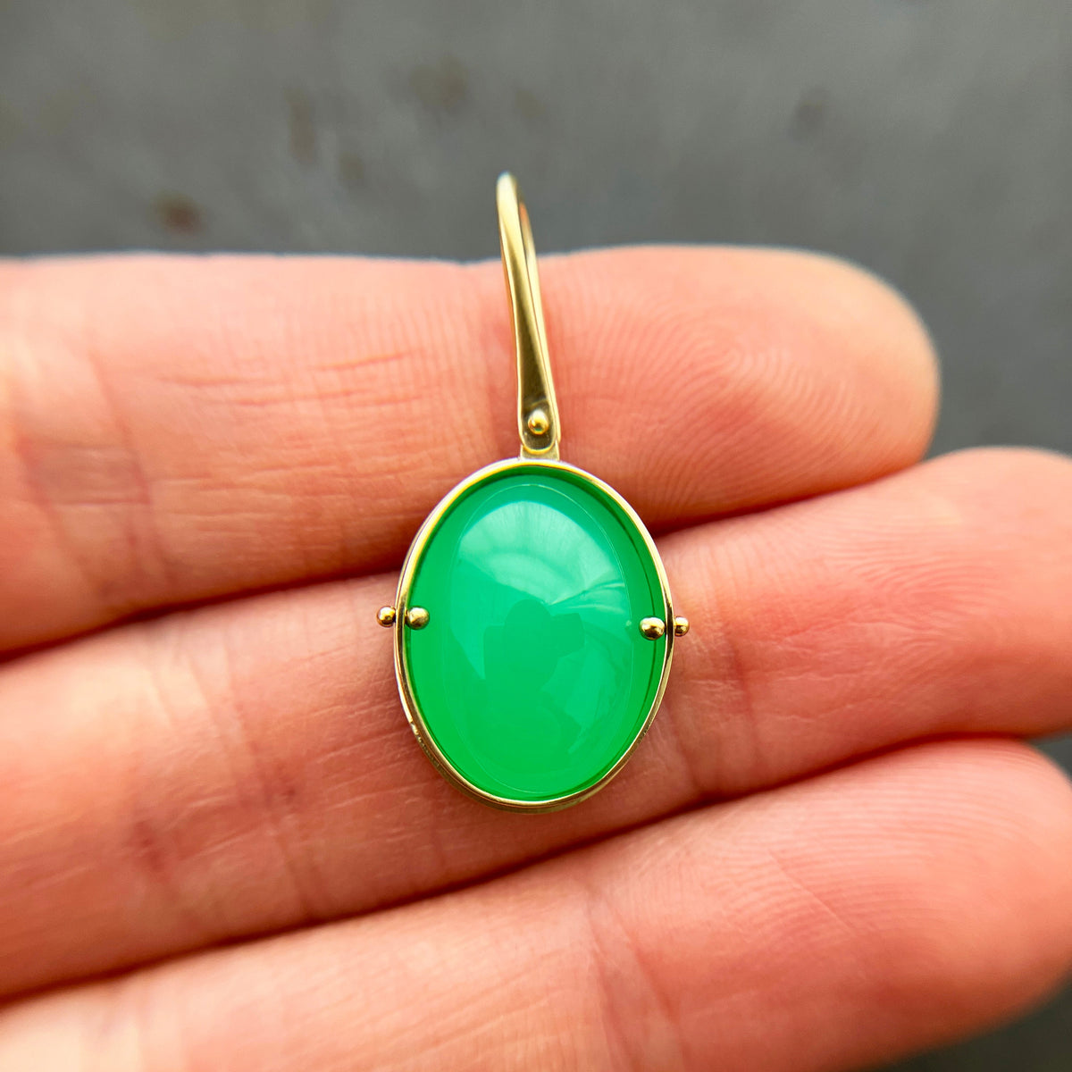 An oval pendant featuring a vibrant green chrysoprase gemstone in a gold bezel setting with a gold hook, displayed on a hand.
