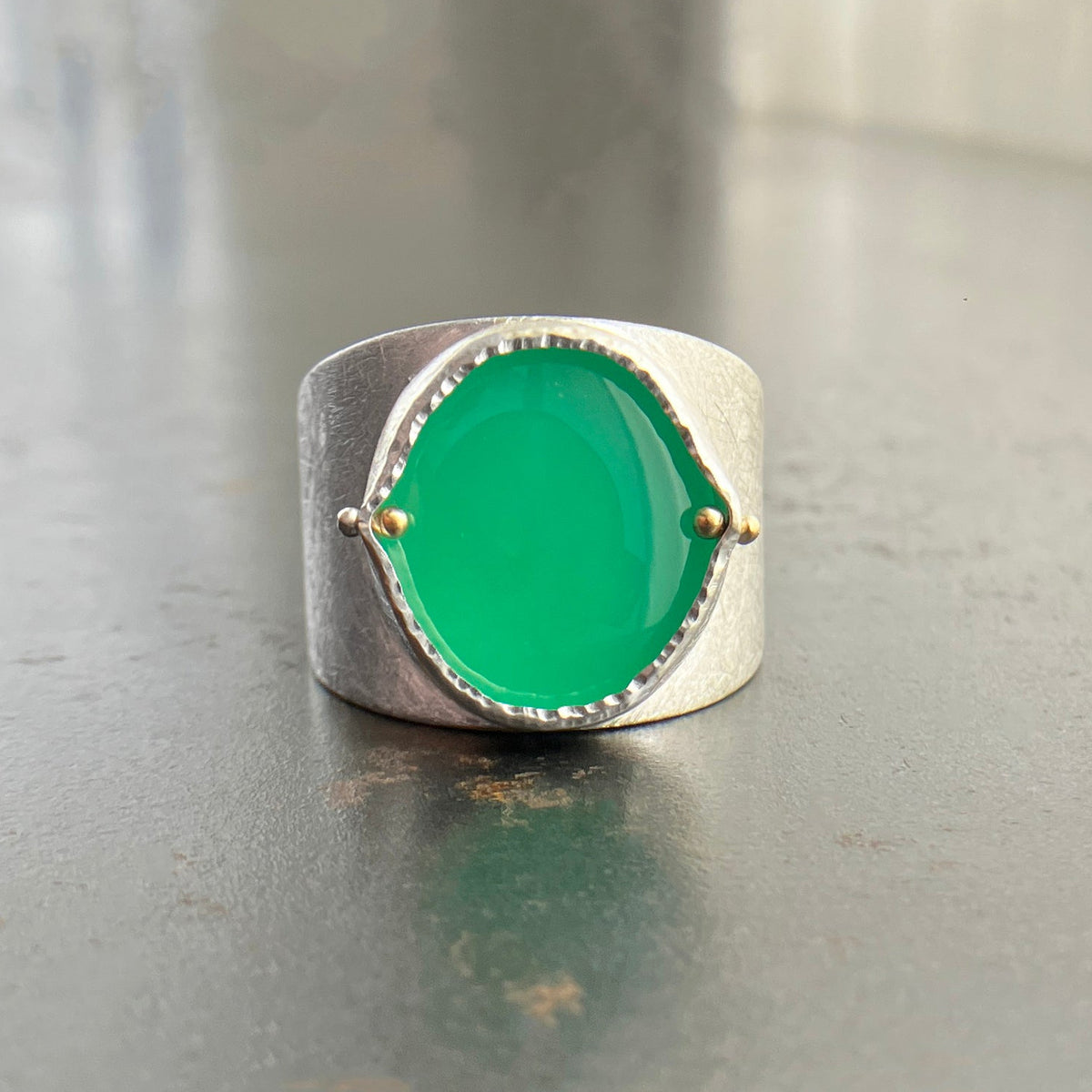 A wide silver band ring featuring a large, polished green chrysoprase teardrop-shaped gemstone in a ball rivet setting with small gold accents, resting on a textured gray surface.