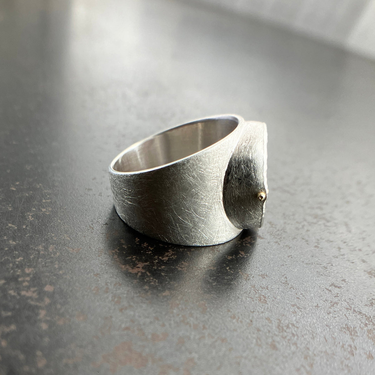 A wide silver band ring featuring a large, polished green chrysoprase teardrop-shaped gemstone in a ball rivet setting with small gold accents, resting on a textured gray surface.