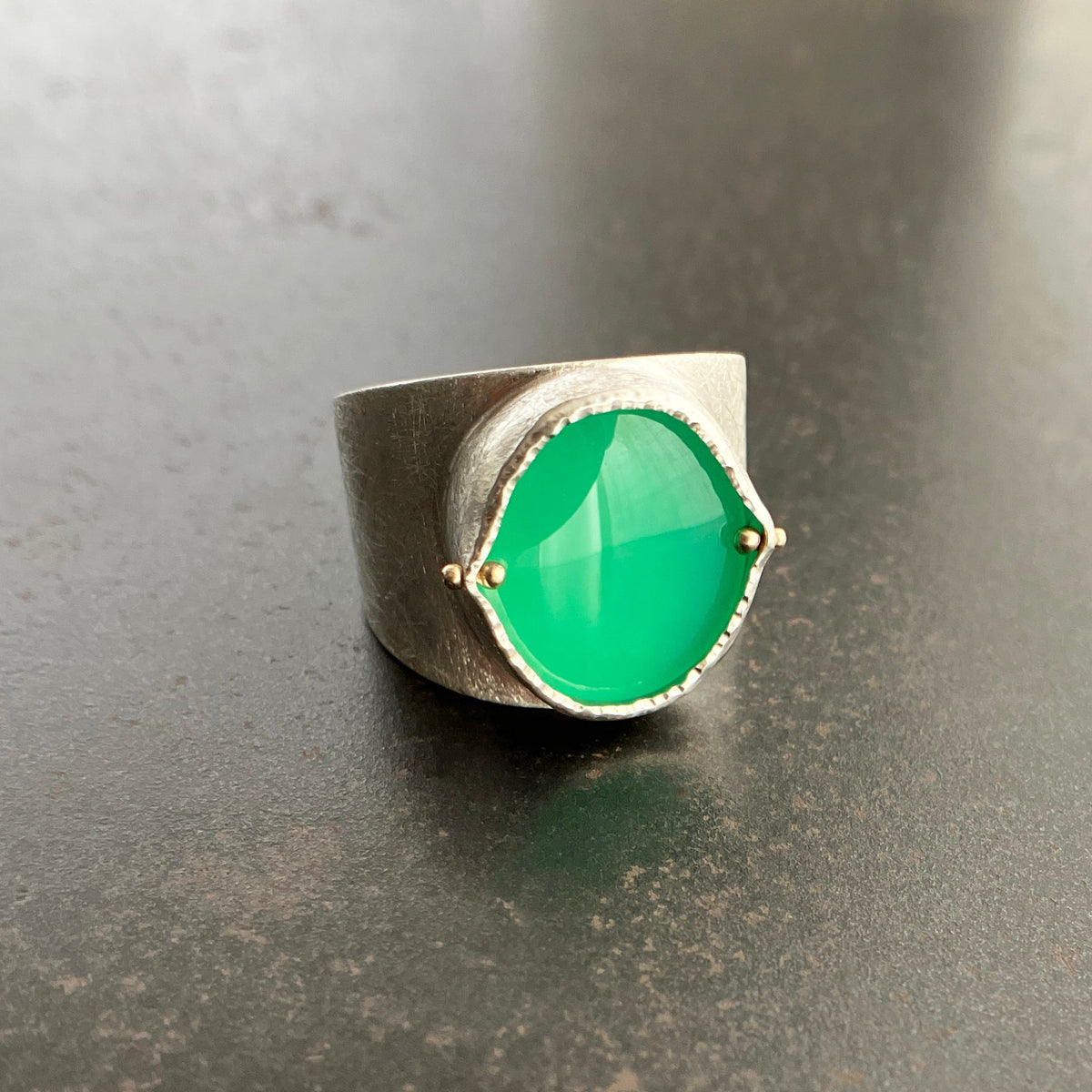 A wide silver band ring featuring a large, polished green chrysoprase teardrop-shaped gemstone in a ball rivet setting with small gold accents, resting on a textured gray surface.
