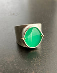 A wide silver band ring featuring a large, polished green chrysoprase teardrop-shaped gemstone in a ball rivet setting with small gold accents, resting on a textured gray surface.