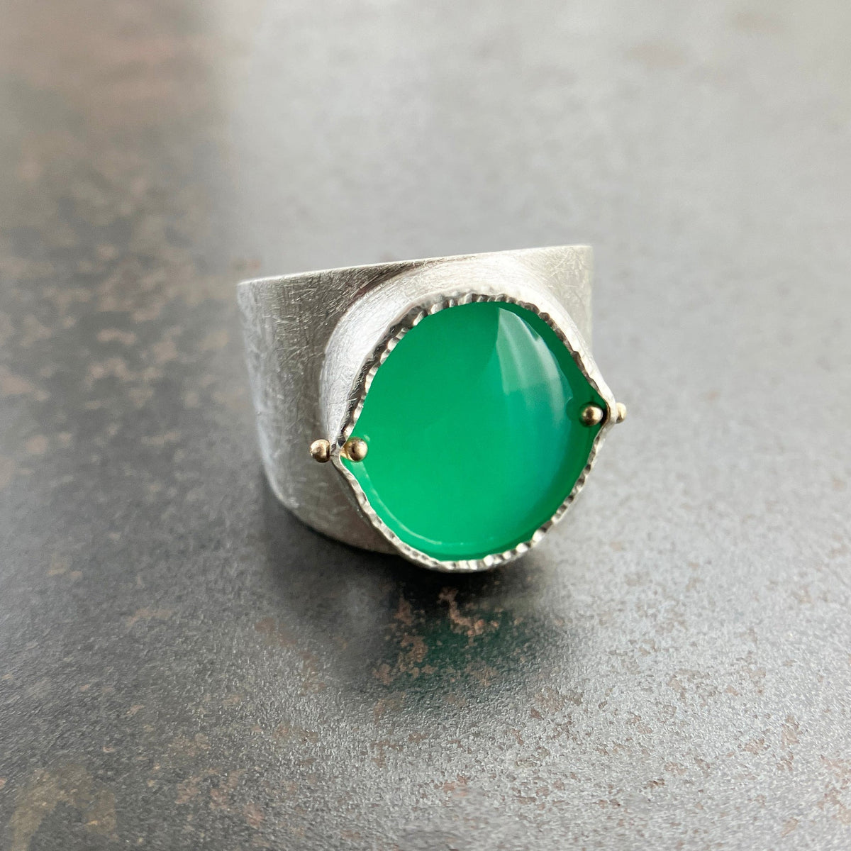 A wide silver band ring featuring a large, polished green chrysoprase teardrop-shaped gemstone in a ball rivet setting with small gold accents, resting on a textured gray surface.