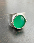 A wide silver band ring featuring a large, polished green chrysoprase teardrop-shaped gemstone in a ball rivet setting with small gold accents, resting on a textured gray surface.