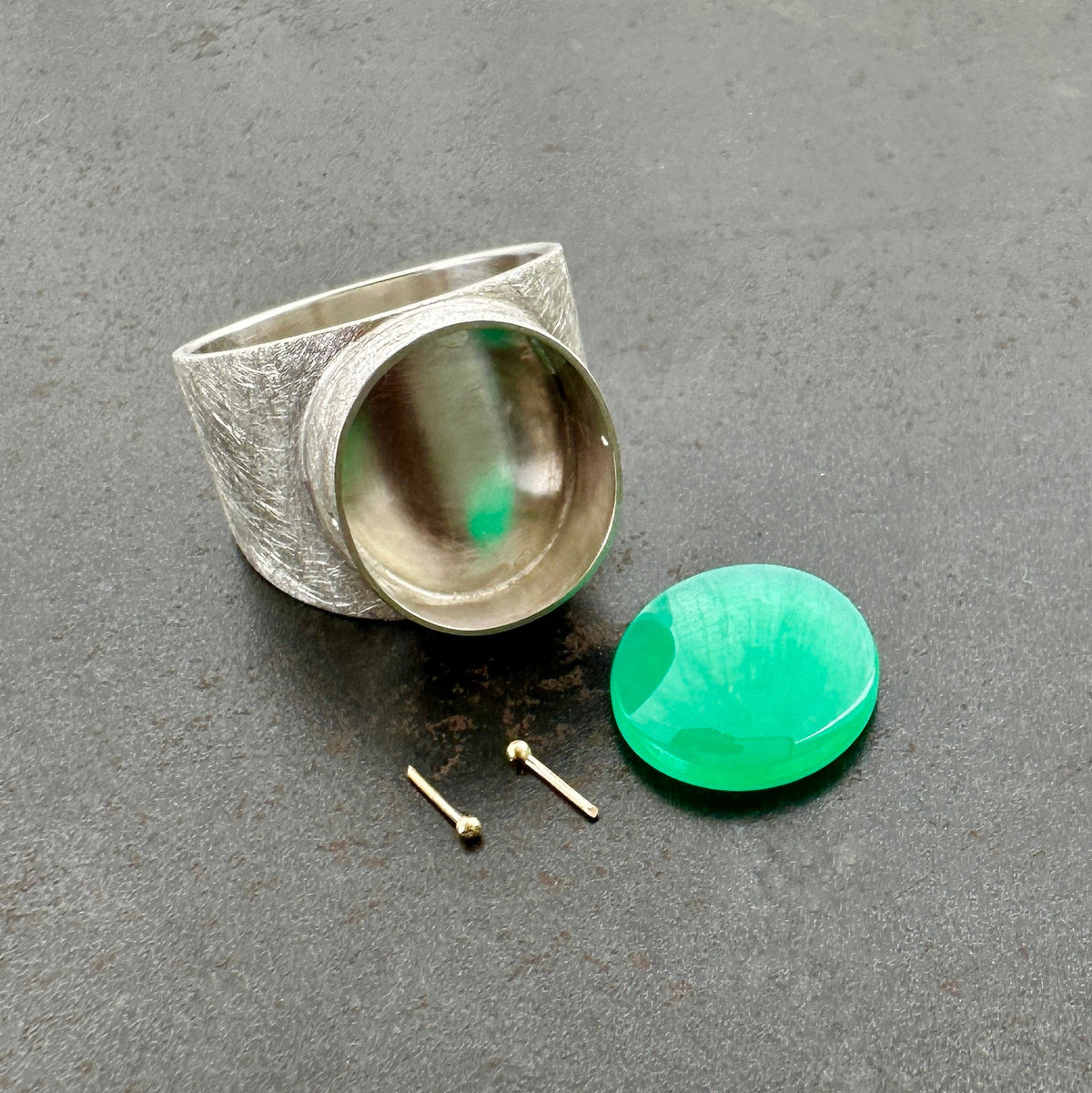 A wide silver band ring featuring a large, polished green chrysoprase teardrop-shaped gemstone in a ball rivet setting with small gold accents, resting on a textured gray surface.