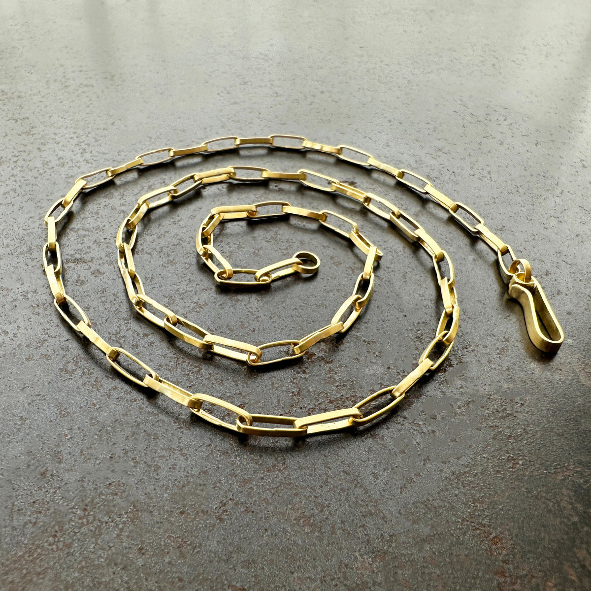 A gold elongated oval chain necklace coiled in a spiral on a textured gray surface.
