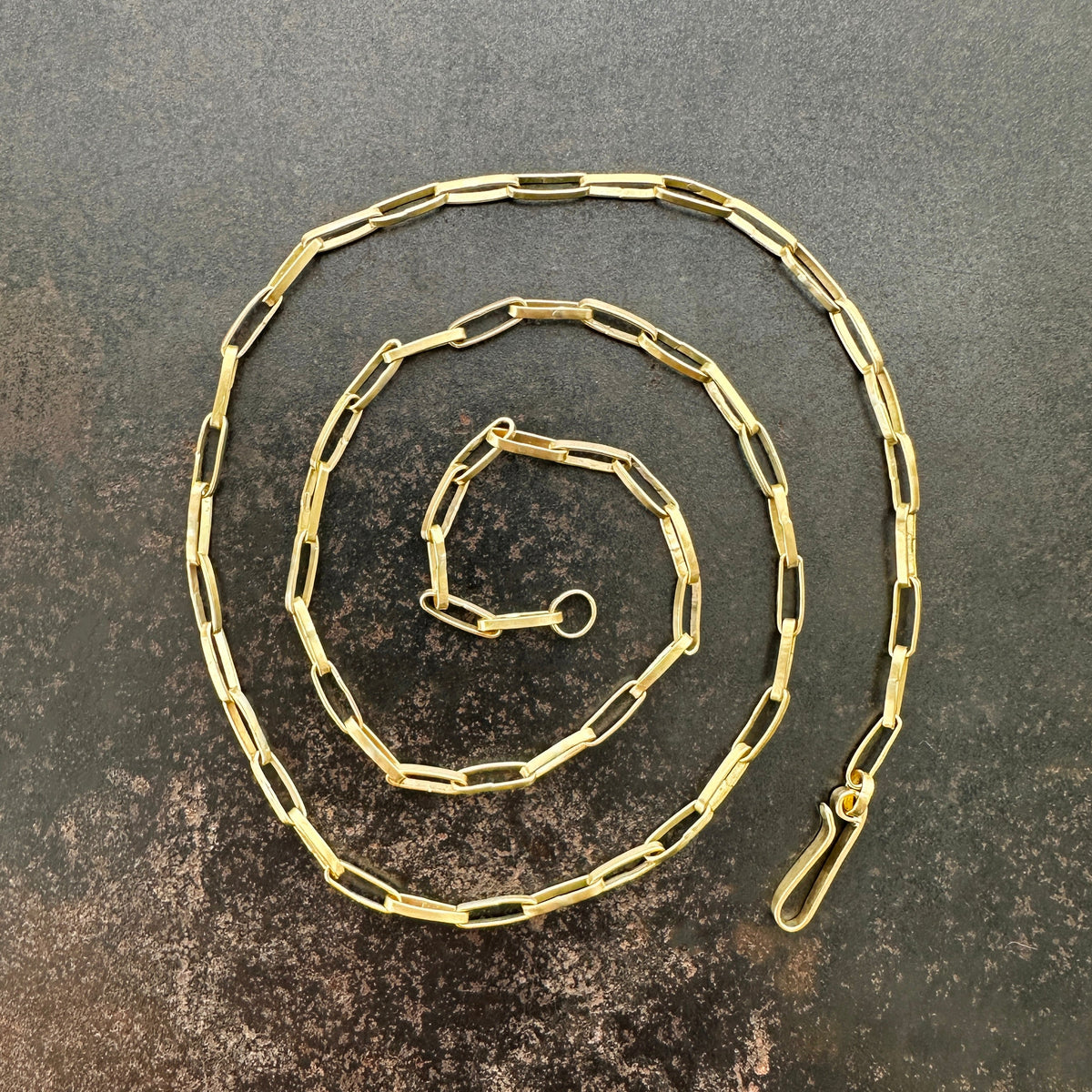 A gold elongated oval chain necklace coiled in a spiral on a textured gray surface.