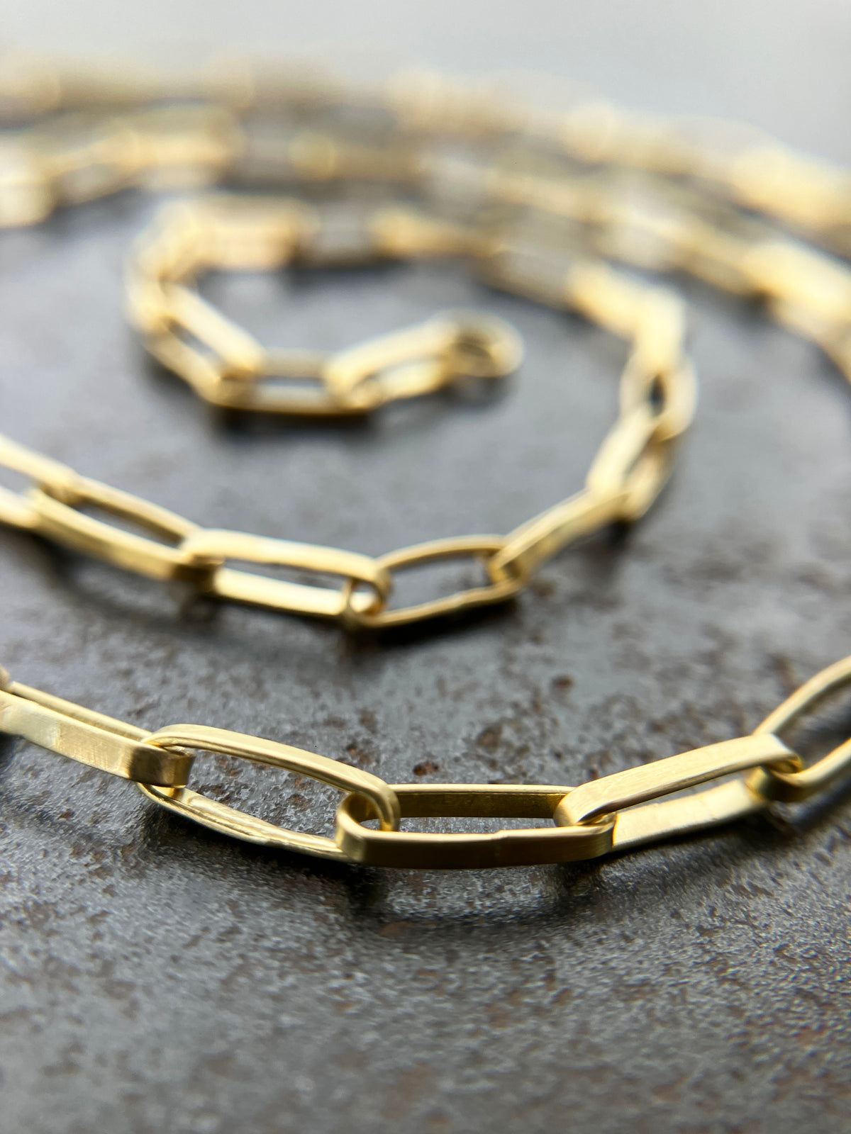 A gold elongated oval chain necklace coiled in a spiral on a textured gray surface.