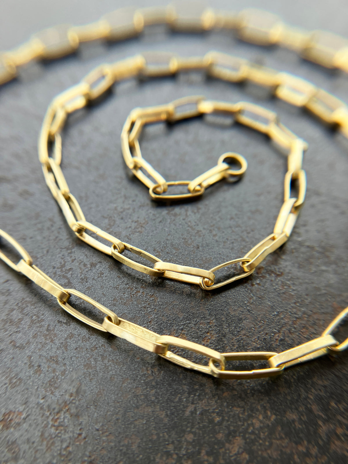 An 18k gold elongated oval chain necklace coiled in a spiral on a textured gray surface.