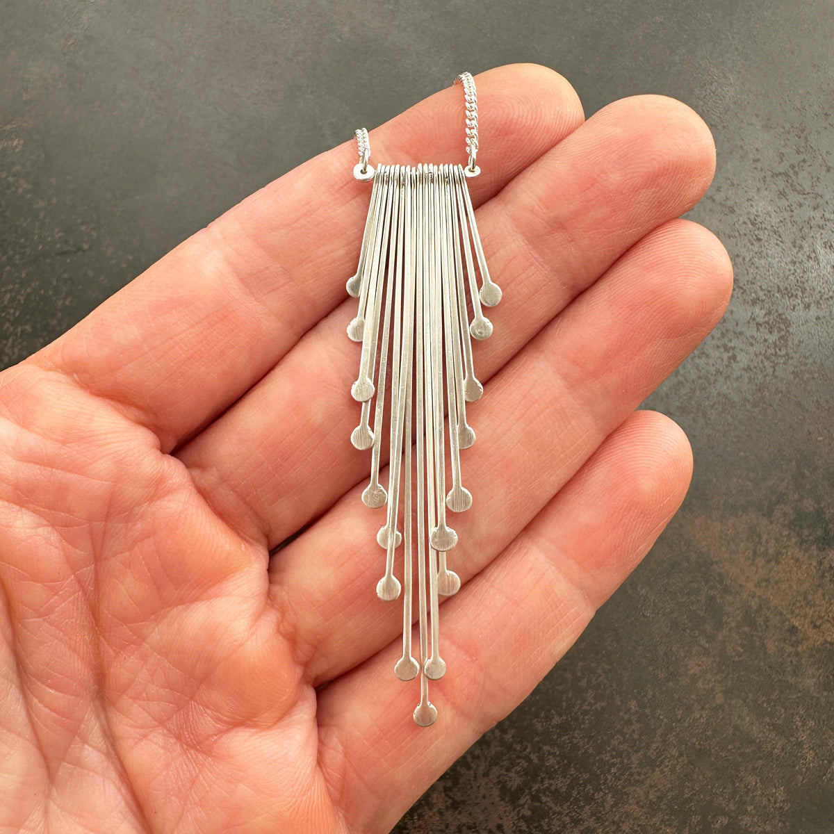 Silver fringe necklace held in a hand against a dark background