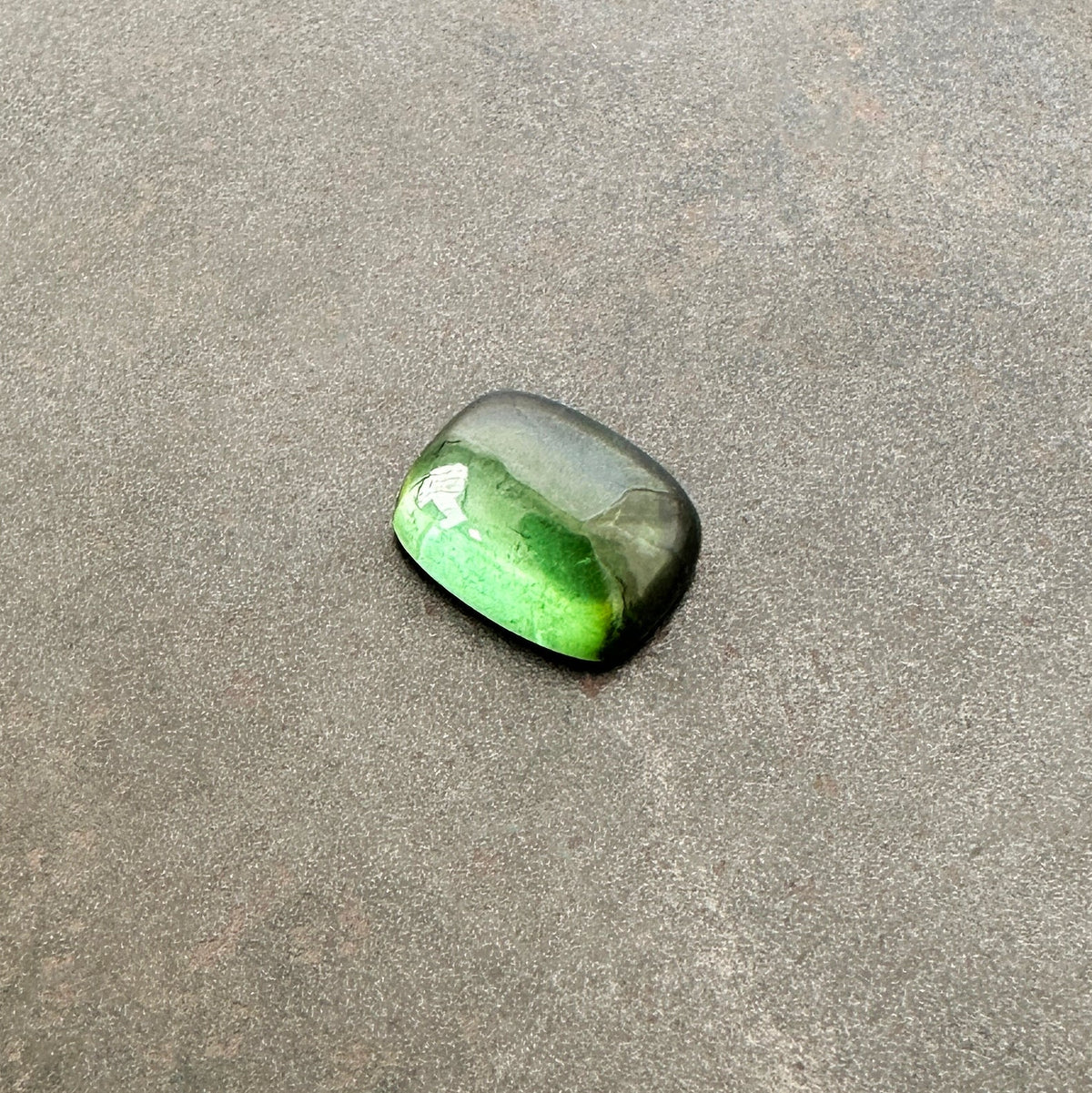 A polished green tourmaline cabochon with a cushion-cut shape, resting on a textured gray stone surface.