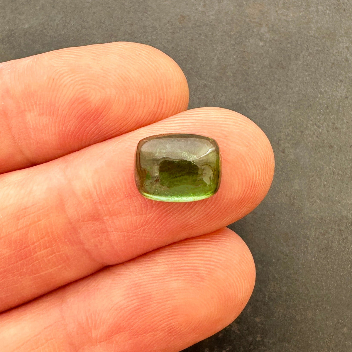 A polished green tourmaline cabochon with a cushion-cut shape, resting on a textured gray stone surface.