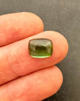 A polished green tourmaline cabochon with a cushion-cut shape, resting on a textured gray stone surface.