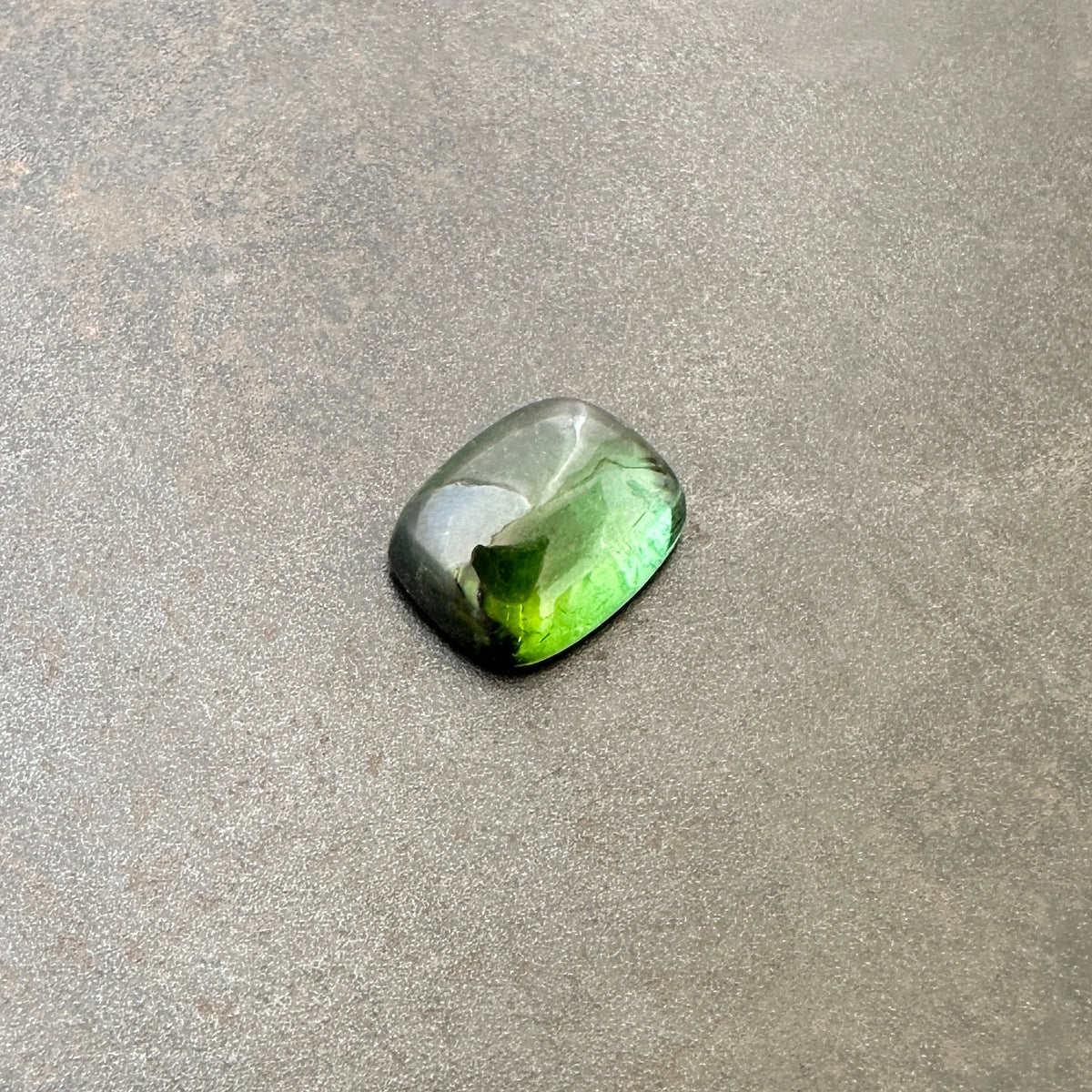A polished green tourmaline cabochon with a cushion-cut shape, resting on a textured gray stone surface.