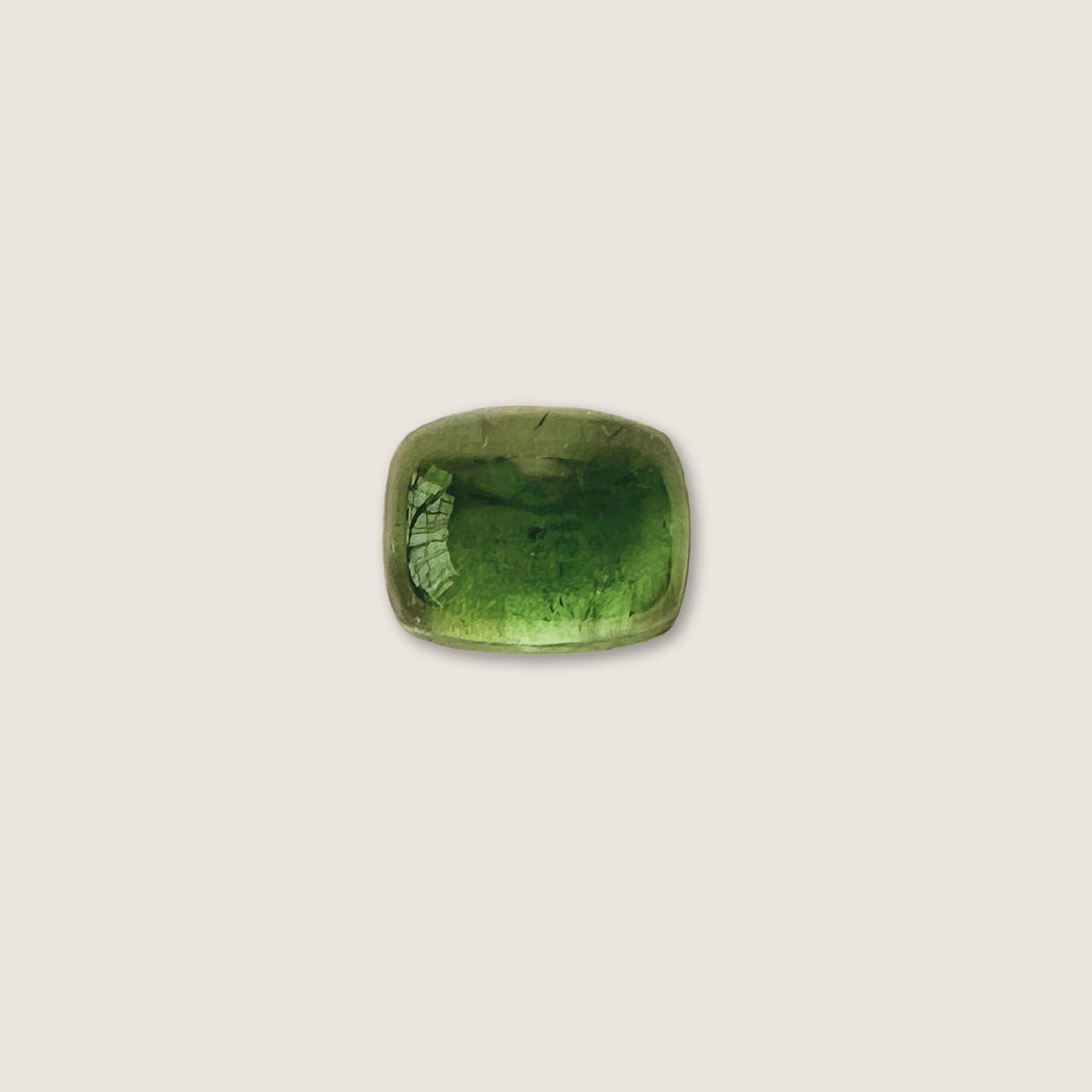 A polished green tourmaline cabochon with a cushion-cut shape, resting on a textured gray stone surface.