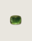 A polished green tourmaline cabochon with a cushion-cut shape, resting on a textured gray stone surface.