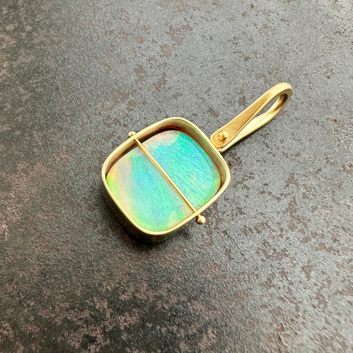 A square pendant with a gold-toned frame and loop, featuring a vibrant iridescent green-blue opal stone, resting on a textured dark gray surface.
