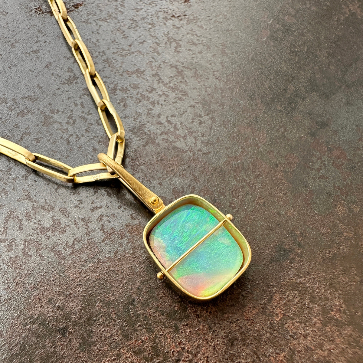 A gold pendant necklace featuring a square iridescent opal stone with green, blue, and pink hues, set in a gold frame and suspended from a gold chain on a textured dark brown surface.