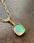 A gold pendant necklace featuring a square iridescent opal stone with green, blue, and pink hues, set in a gold frame and suspended from a gold chain on a textured dark brown surface.
