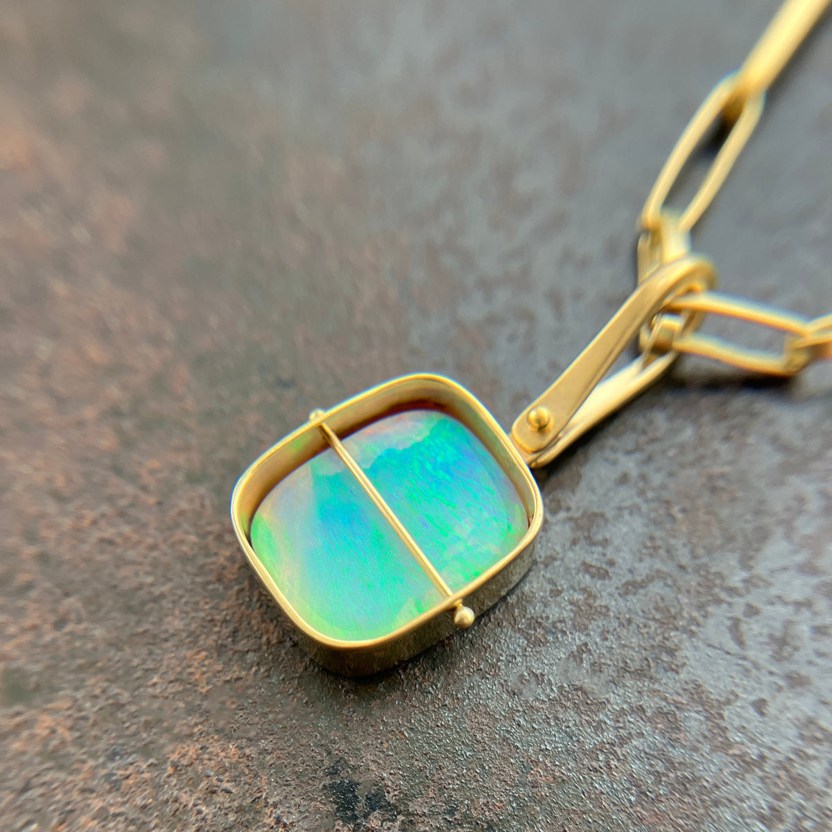 A gold pendant necklace featuring a square iridescent opal stone with green and blue play-of-color, set in a gold bezel with a central gold bar, resting on a textured dark gray surface.