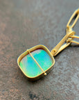 A gold pendant necklace featuring a square iridescent opal stone with green and blue play-of-color, set in a gold bezel with a central gold bar, resting on a textured dark gray surface.