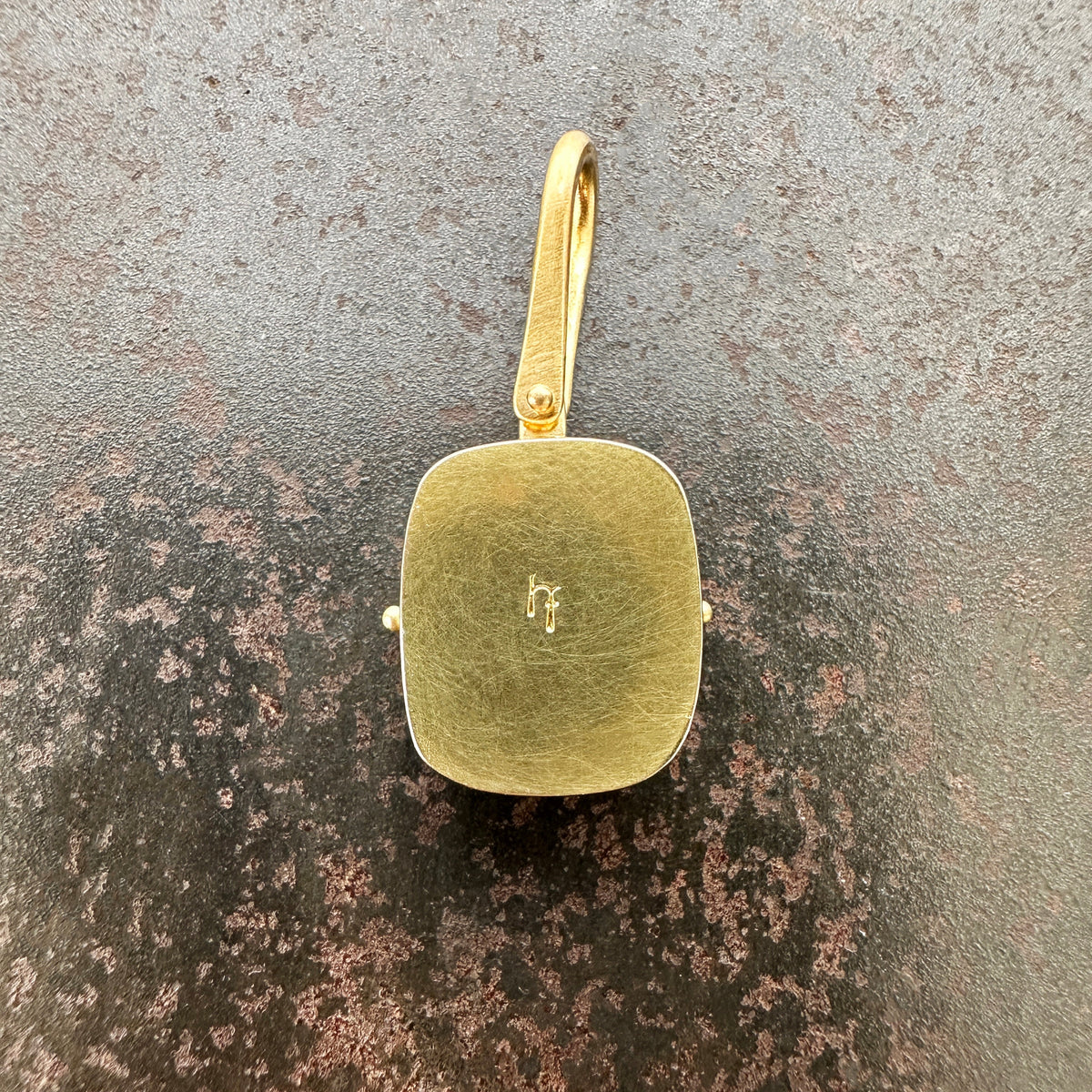 A gold pendant with a rounded rectangular cushion-cut face and a top loop, displayed on a textured dark gray and brown surface.