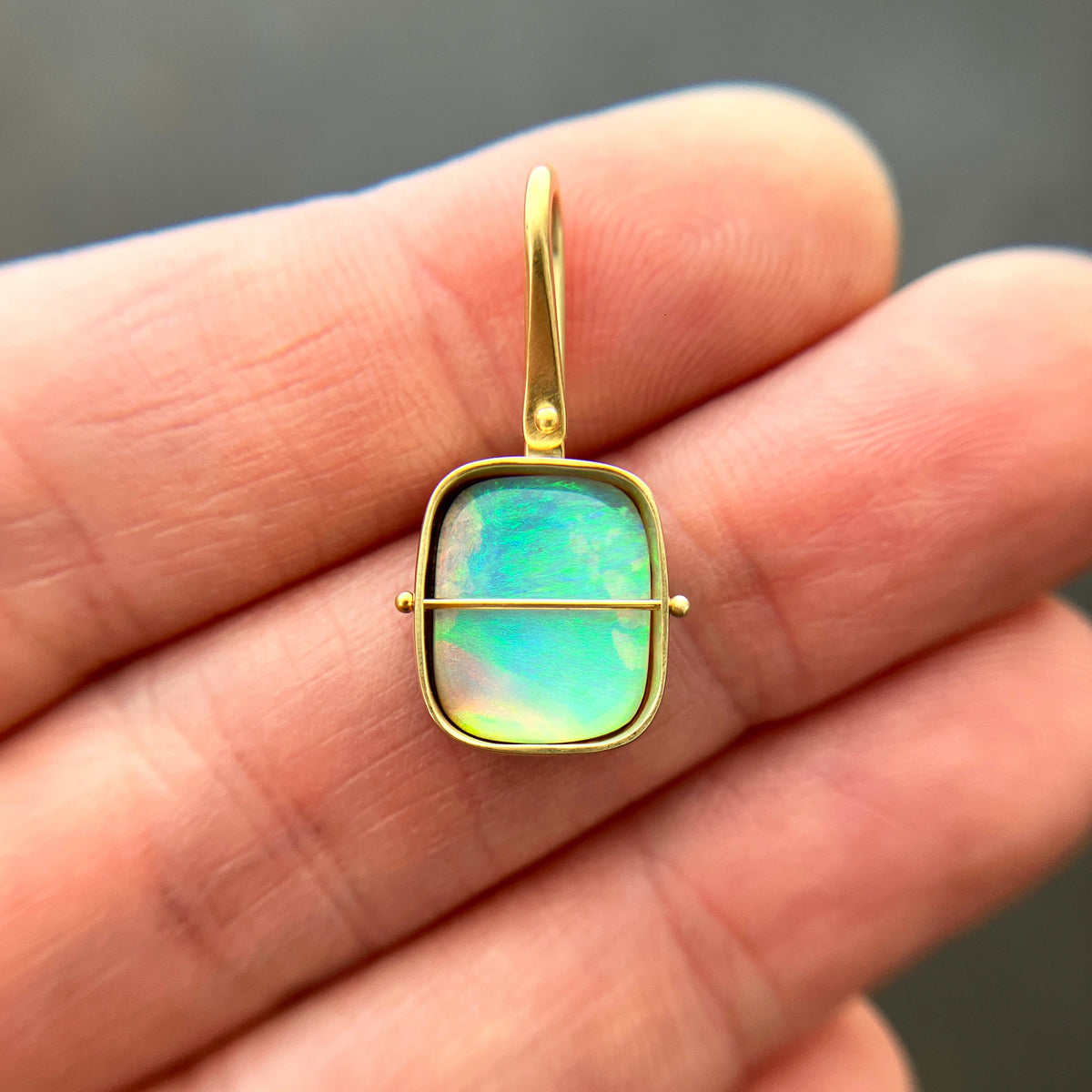 A gold pendant with a square iridescent opal stone held in a person's hand. The pendant features a gold bezel and loop, with a thin horizontal gold bar across the center.