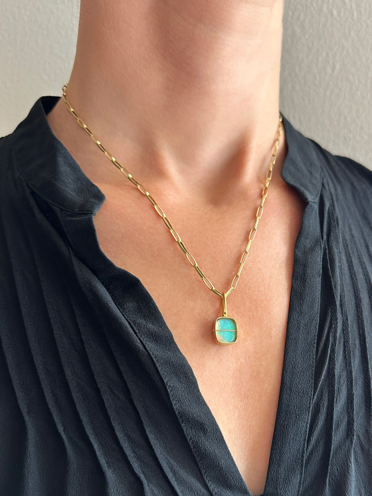 A gold pendant with a square iridescent opal stone held in a person's hand. The pendant features a gold bezel and loop, with a thin horizontal gold bar across the center.