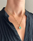 A gold pendant with a square iridescent opal stone held in a person's hand. The pendant features a gold bezel and loop, with a thin horizontal gold bar across the center.