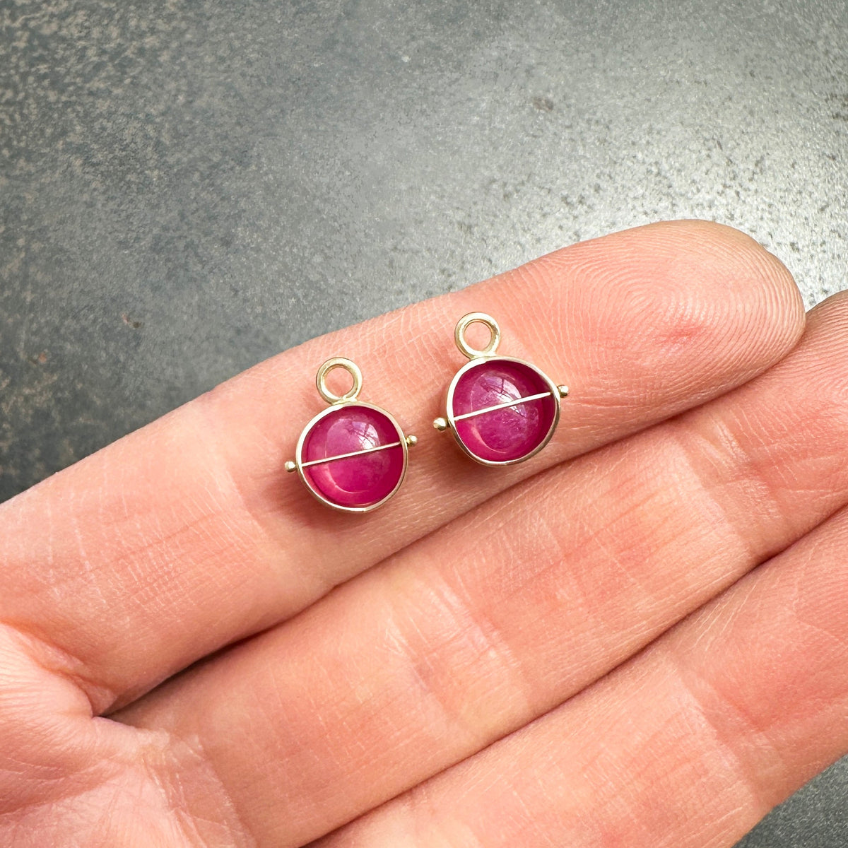 A pair of small 14k gold charm hoop earrings with round ruby gemstones, shown resting on an open hand against a gray textured background.