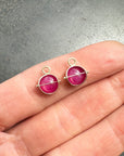 A pair of small 14k gold charm hoop earrings with round ruby gemstones, shown resting on an open hand against a gray textured background.