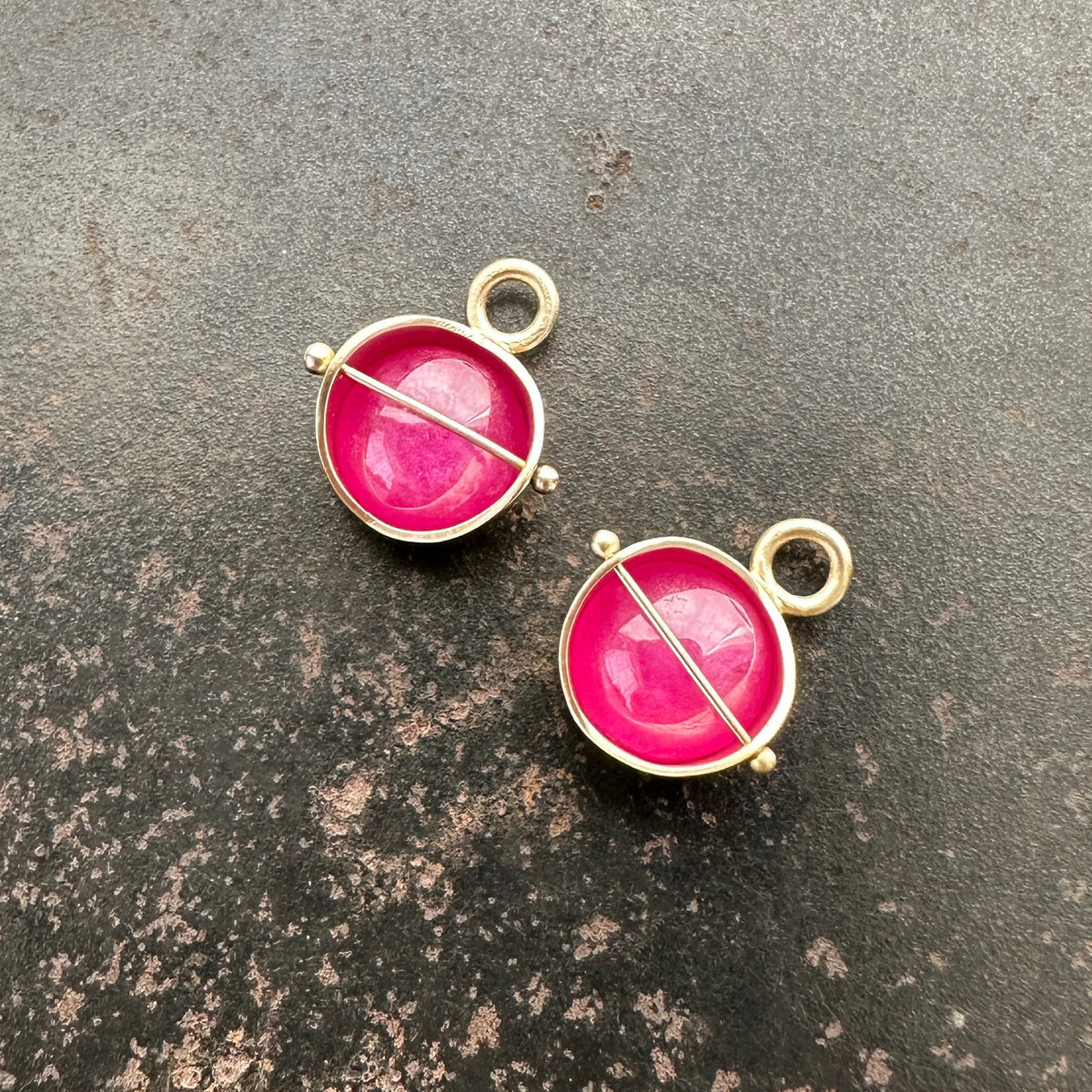 Two 14k gold hoop charms with vibrant pink ruby cabochon gemstones and a central gold bar, displayed on a dark textured surface.