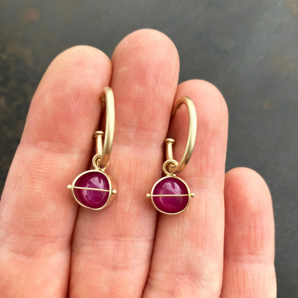 A pair of small 14k gold hoop earrings with ruby gemstones, held between a person's fingers against a dark gray background.