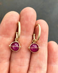 A pair of small 14k gold hoop earrings with ruby gemstones, held between a person's fingers against a dark gray background.