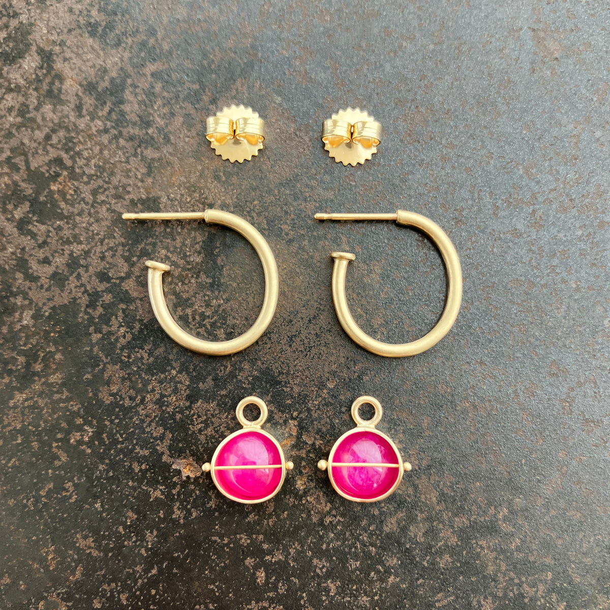 A set of 14k gold hoop earrings laid flat on a textured dark gray surface, including two small round ruby hoop charm earrings, two open hoop earrings, and two drop earrings with bright pink oval stones in gold settings.