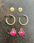 A set of 14k gold hoop earrings laid flat on a textured dark gray surface, including two small round ruby hoop charm earrings, two open hoop earrings, and two drop earrings with bright pink oval stones in gold settings.