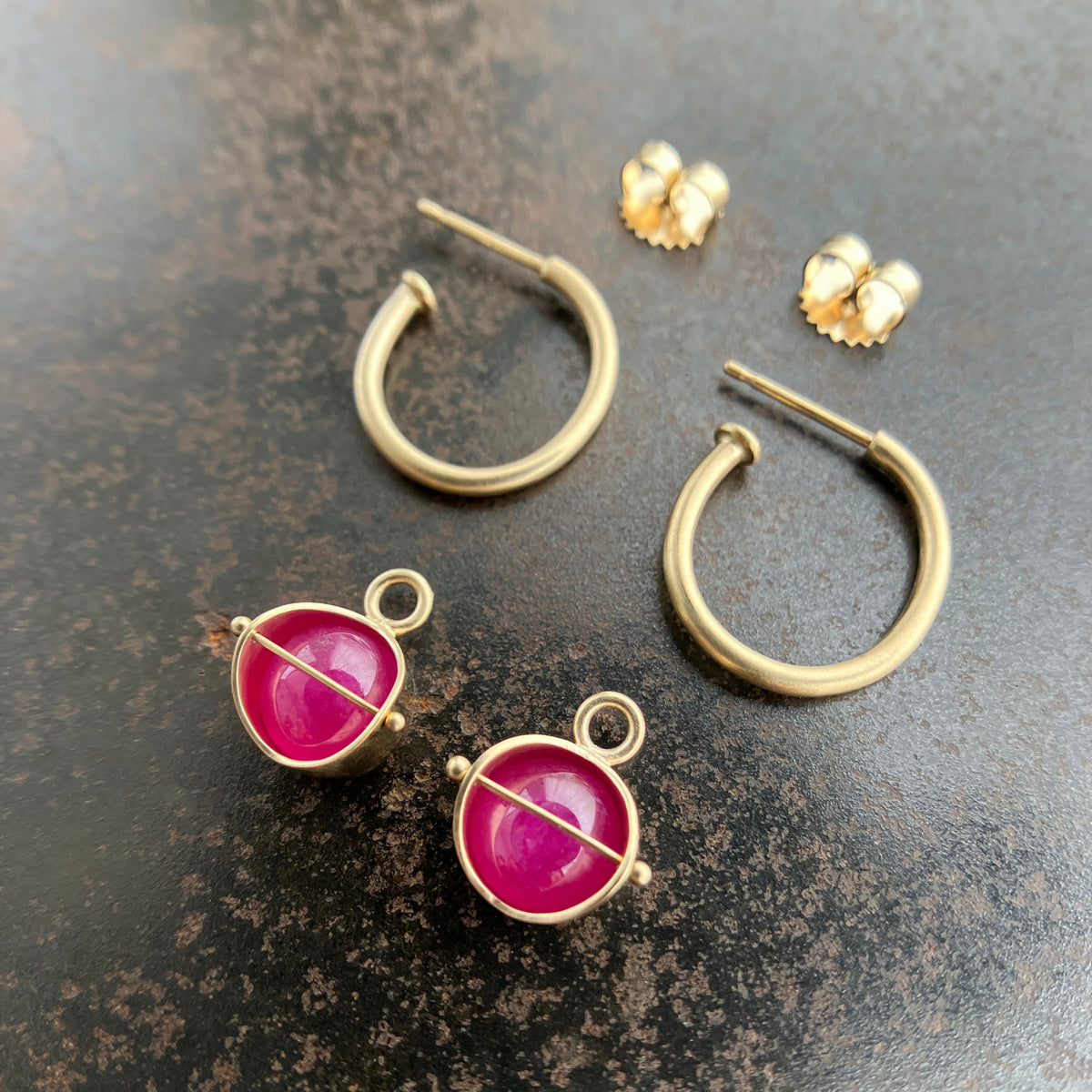 A pair of 14k gold hoop earrings with round magenta-pink ruby gemstone hoop charms displayed on a dark textured surface.