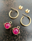 A pair of 14k gold hoop earrings with round magenta-pink ruby gemstone hoop charms displayed on a dark textured surface.