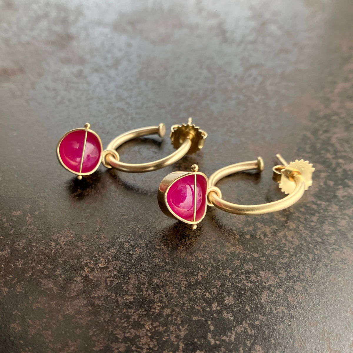 A pair of gold hoop earrings with pink ruby cabochon gemstone hoop charms, displayed on a dark textured surface.